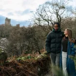 Scenic woodland walk with two people in winter coats, passing a stone castle partially hidden by trees on a hillside.