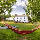 Colourful hammock strung between two trees in a grassy garden, with a white farmhouse and trampoline in the background under a blue, partly cloudy sky