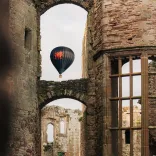 Hot air balloon with a red dragon design floating in the sky, framed by the stone arches and windows of a historic castle ruin.
