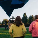 Three people standing on grass watching a hot air balloon being prepared for flight, with trees and a clear sky in the background.