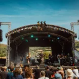 band playing at festival on a sunny day with crowd watching and hills in background.