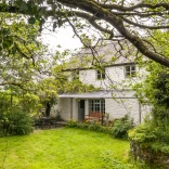 A white painted stone cottage, with a seating area outside, in a lawned garden.