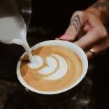 Here’s concise alt text for the second image — and as requested, no mention of any blurred face:  **Alt text:**   *A close-up of steamed milk being poured into a cup of coffee to create latte art and hand with tattoo and ring.