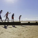 A family walking along a wooden groyne on a sandy beach, with bright sunlight and a clear sky in the background.