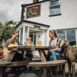 Family sit at a wooden picnic table outside a white pub with drinks, while two dogs stand nearby.
