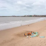 Surf lesson group with surfboards gathered on a wide sandy beach, with gentle waves and a coastal town in the distance.