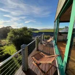 A wooden balcony with folding deckchairs overlooking green countryside, with sunlight streaming across the landscape and large glass doors opening into a modern dining and kitchen area.