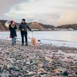 Couple and dog on a pebble beach beside a calm bay, with distant coastal village and hills under a cloudy sky.