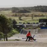 A child in an adapted sled / ski-pram being pushed by a man on a dry ski-slope.