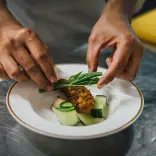 Close‑up of a chef's hands garnishing a plated dish with fresh herbs and sliced vegetables.
