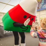 A costumed mascot in red and green leans down to greet a small child at an indoor event.