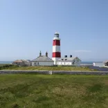 Red and white lighthouse and small buildings on a low stone‑walled headland with ocean views and clear blue sky.