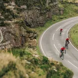 Cyclists climbing a winding mountain road past a rock face painted with a Welsh dragon and the words “Cymru am byth” and “...the Valleys”, surrounded by green hills.