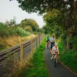 Adult and child cycling along a narrow path beside railway tracks, with two more riders following under trees.