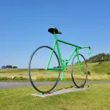 Green oversized bicycle sculpture standing beside parkland road in Pembrey Country Park.