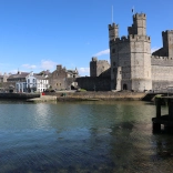Castell Caernarfon wrth lan y dŵr, gyda’i furiau cerrig mawreddog yn cael eu hadlewyrchu yn nyfroedd tawel yr harbwr o dan awyr las.