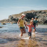 Family paddling and splashing in shallow water at a rocky cove on a sunny day, with kayaks visible offshore and rugged coastal cliffs in the background.