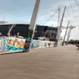 Colourful street art murals lining the Riverside Walk beside the Principality Stadium in Cardiff, with the stadium’s distinctive support masts and the River Taff alongside.