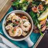 Bowl of prawns in a herb dressing served with mixed leaves, cherry tomatoes and a lemon wedge on a blue plate.