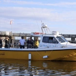 Passengers aboard the Storm Lee Junior charter boat on calm waters beside a harbour pontoon, with Cardiff Bay visible in the background under a partly cloudy sky.
