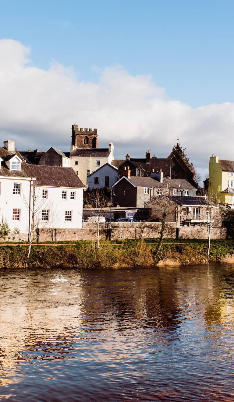River with houses along the riverbank, and the top of Brecon Cathedral in the distance