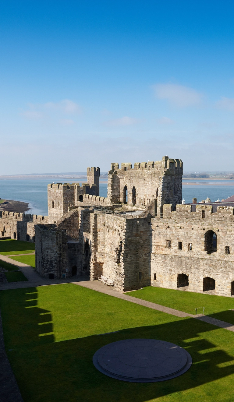 Blick in eine große steinerne Burg, deren Türme Schatten auf den grasbewachsenen Innenhof werfen, mit dem Meer im Hintergrund.