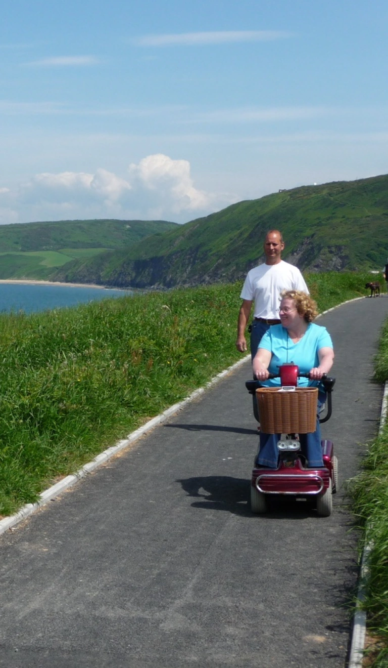 Lady on a mobility scooter with male companion on a tarmac section of the wales coast path looking out to sea
