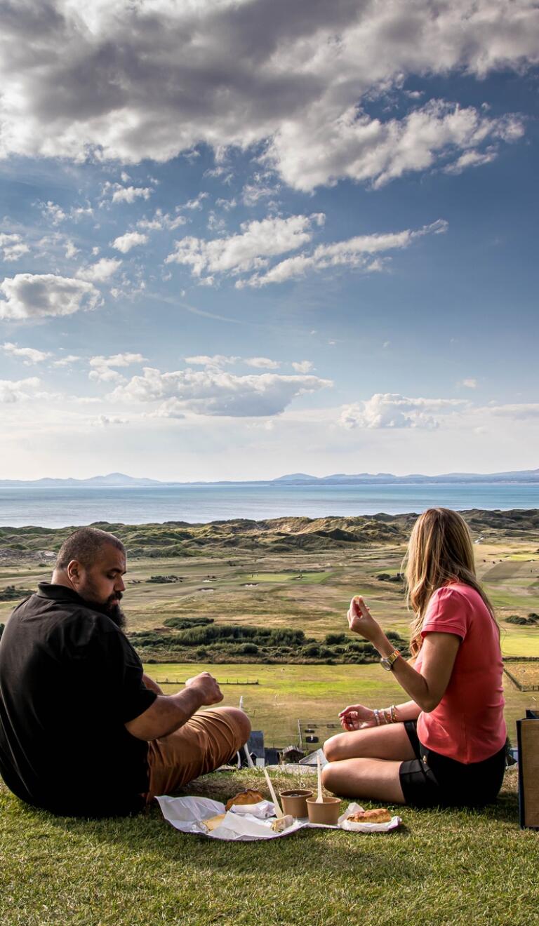 Two people having a picnic overlooking a coastal golf course.
