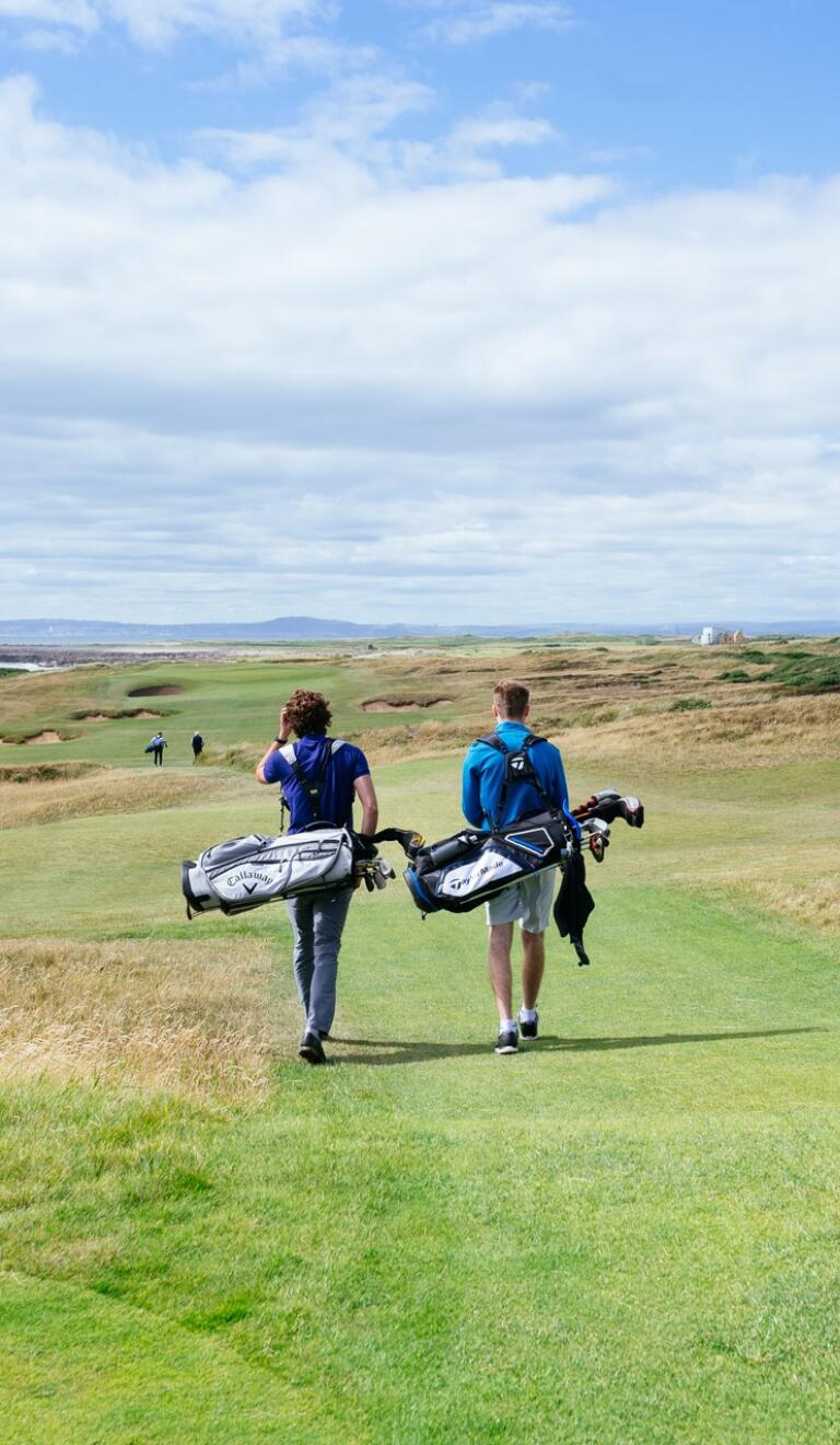 Two people with golf bags walking down a wide pathway on a coastal golf course.