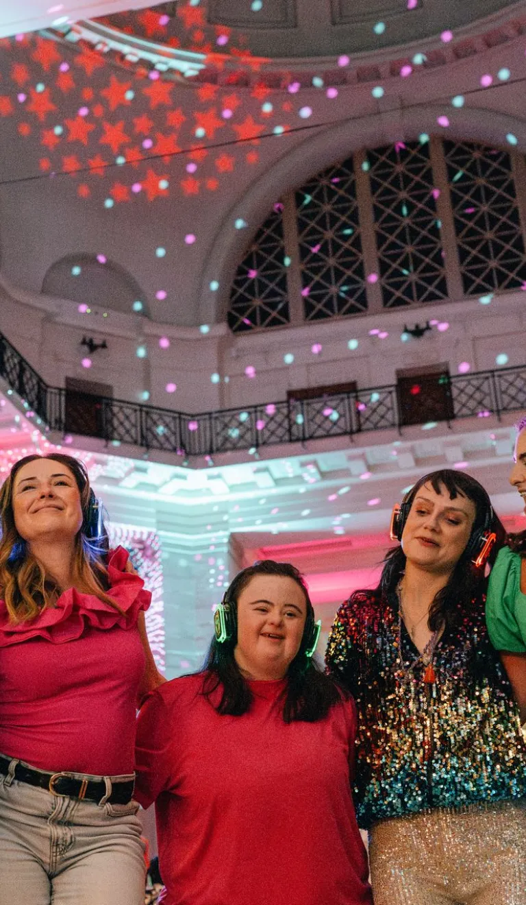 A group of seven friends wearing headphones smiling and dancing at a silent disco in a room with disco lights and a high ceiling.