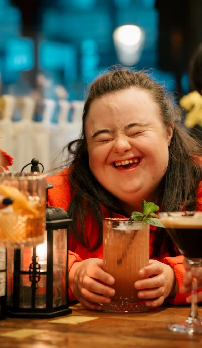 Three people sat around a table with cocktails, the central person is laughing with eyes closed and mouth open.