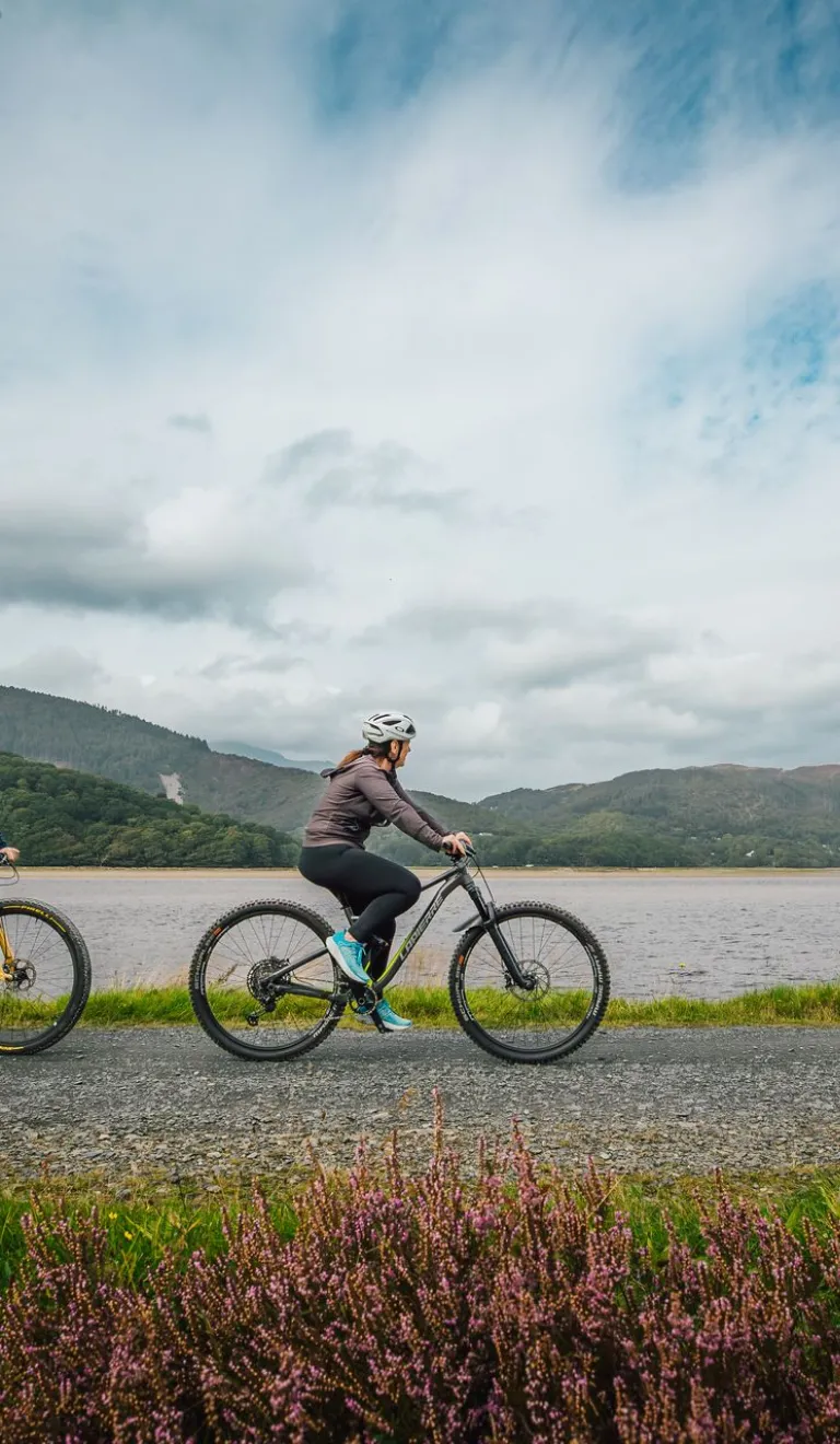 Three cyclists on a path by a river, with mountains in the background.