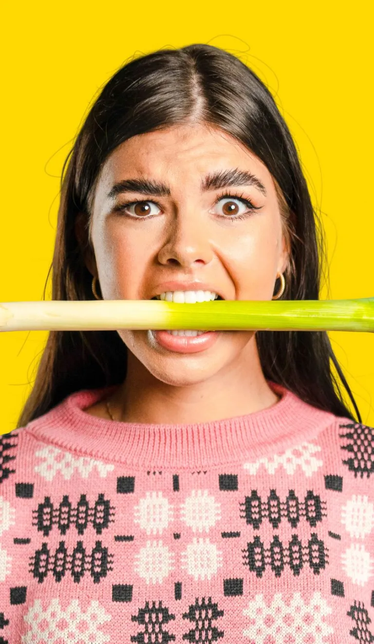 A woman wearing a pink jumper with a leek in her mouth.