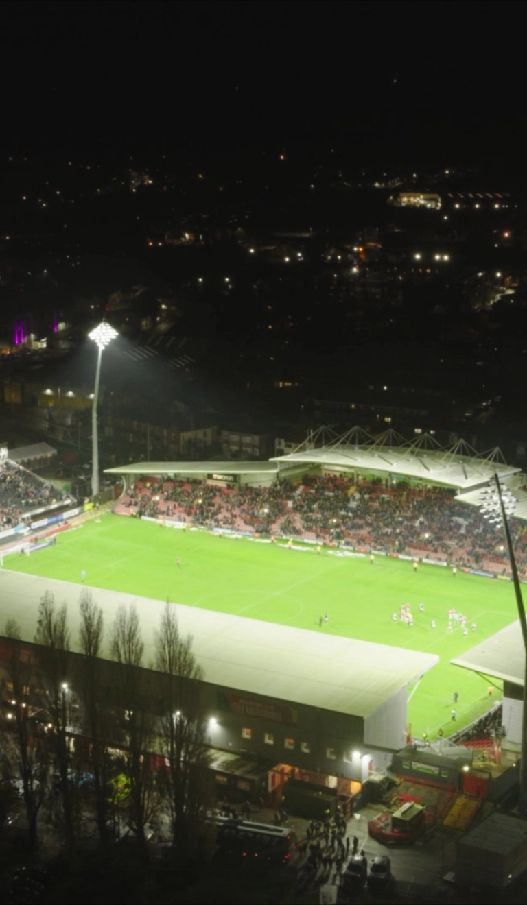 A football stadium in the dark seen from above with flood lights lighting the pitch. THe lights of the surrounding town can be seen in the dark sky around.