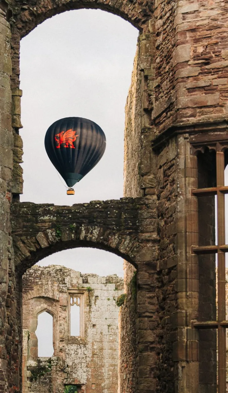 Hot air balloon with a red dragon design floating in the sky, framed by the stone arches and windows of a historic castle ruin.