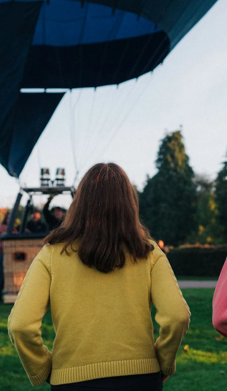 Three people standing on grass watching a hot air balloon being prepared for flight, with trees and a clear sky in the background.