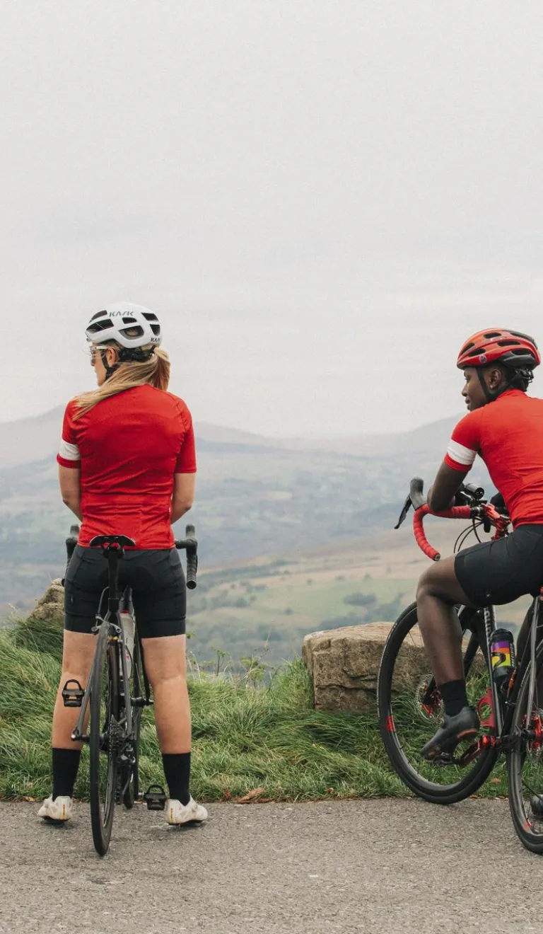 A group of four cyclists wearing red jerseys pause with their bikes at a viewpoint overlooking rolling hills and countryside.