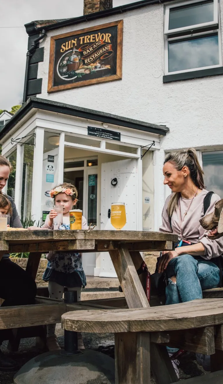 Family sit at a wooden picnic table outside a white pub with drinks, while two dogs stand nearby.