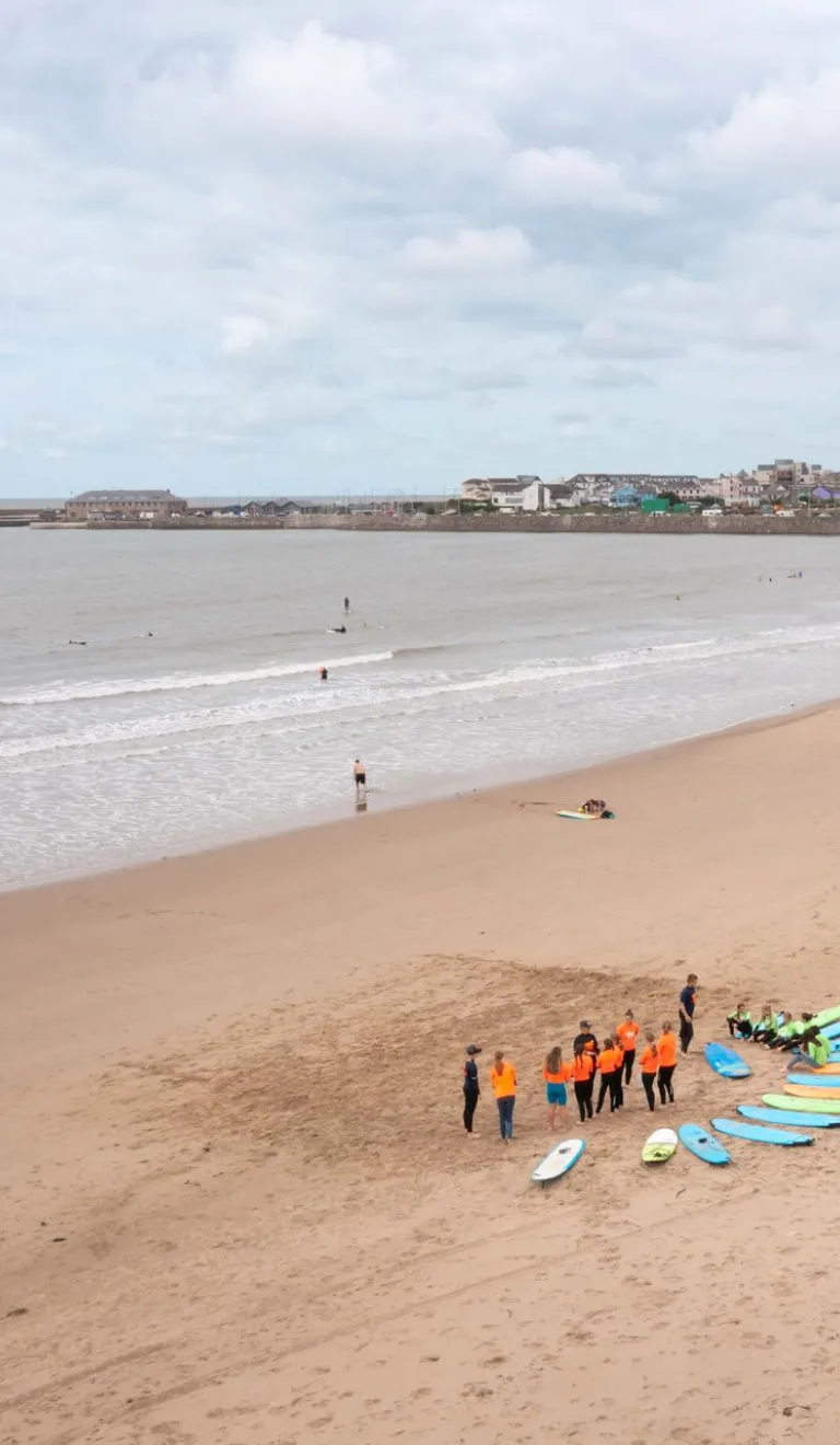 Surf lesson group with surfboards gathered on a wide sandy beach, with gentle waves and a coastal town in the distance.