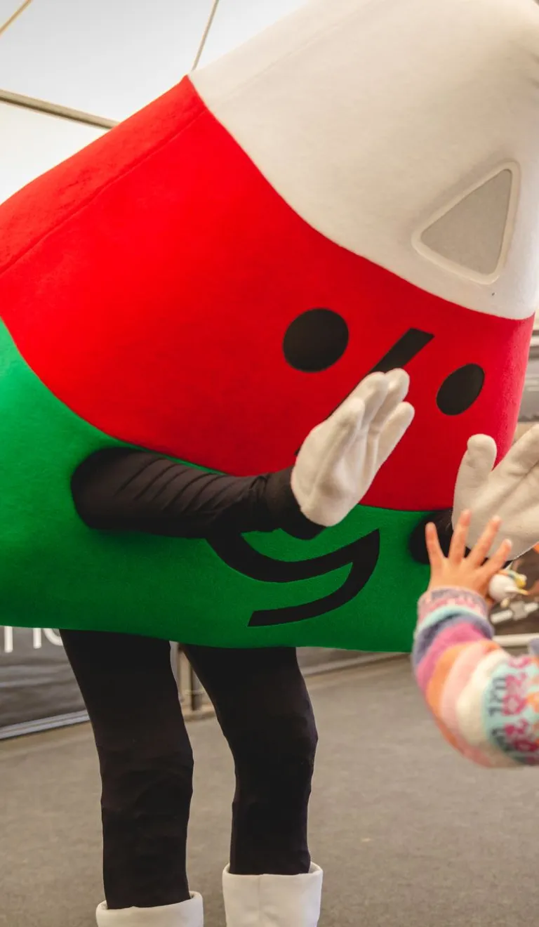 A costumed mascot in red and green leans down to greet a small child at an indoor event.