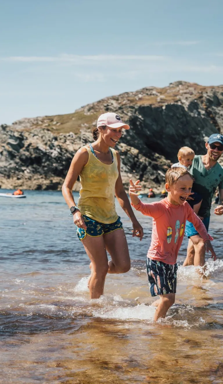 Family paddling and splashing in shallow water at a rocky cove on a sunny day, with kayaks visible offshore and rugged coastal cliffs in the background.