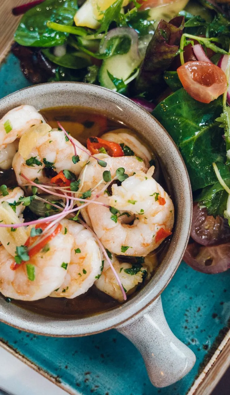 Bowl of prawns in a herb dressing served with mixed leaves, cherry tomatoes and a lemon wedge on a blue plate.