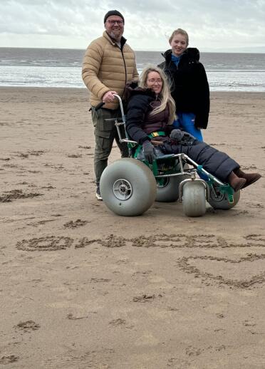 family including woman in beach wheelchair on sandy beach with words @WheelsnoHeels written in sand.