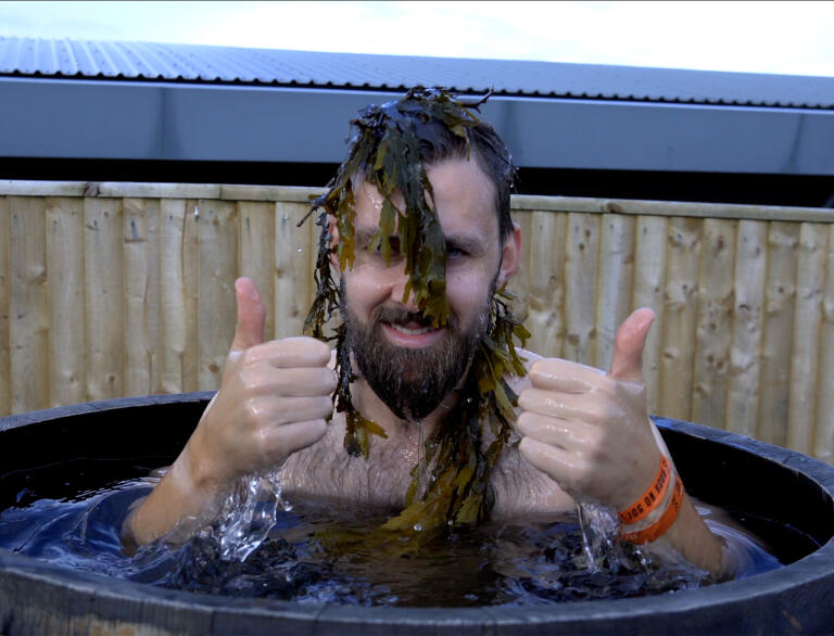 A man in a wooden barrel full of water, with seaweed on his head.