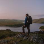 Man with a backpack on his back, stood on a mountain edge looking out across a landscape of mountains and a lake.